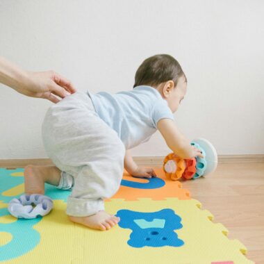 A toddler playing with colorful toys on a puzzle mat indoors, guided by an adult hand.